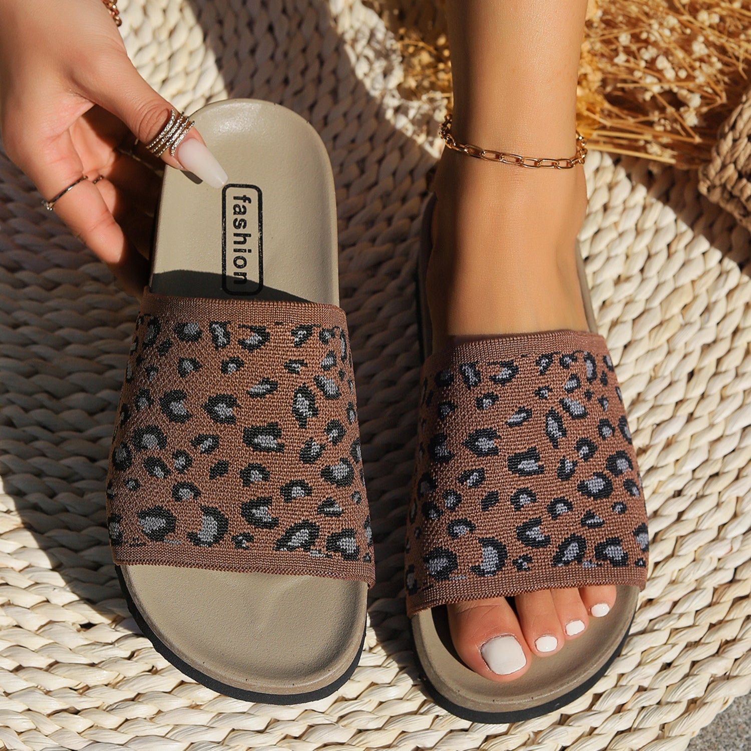 A pair of brown leopard print slide sandals, worn by a woman with a white pedicure.