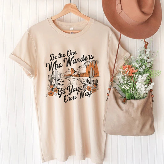 A Western style graphic t shirt with a vintage depiction of a desert road and lettering that says "Be the one who wanders - go your own way."