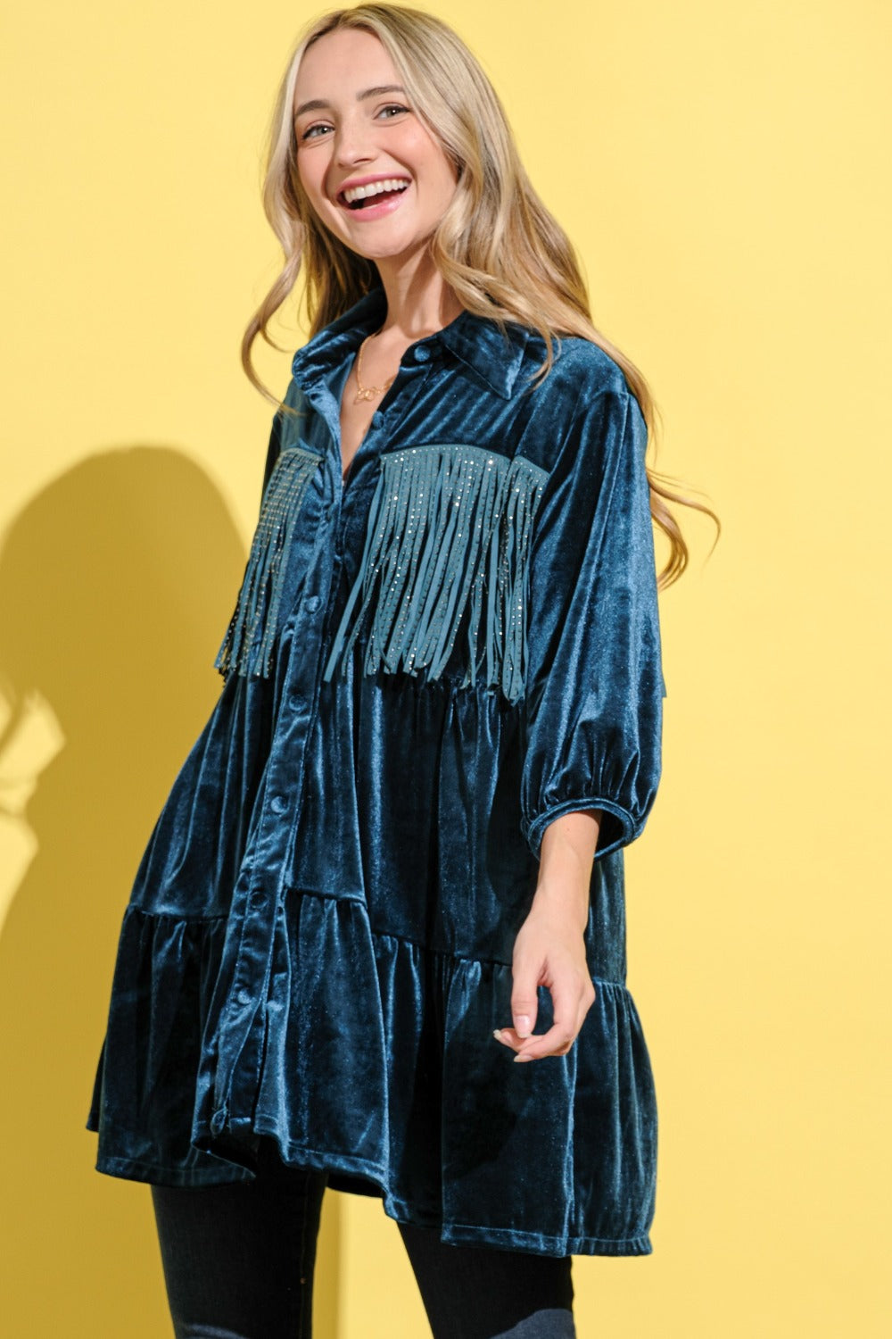 A woman wearing a tiered teal tunic shirt dress embellished with fringe on the front and back. 