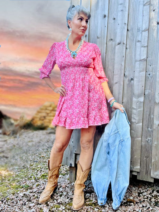 A woman standing against a wooden building wearing a pink floral puff sleeve minidress and cowboy boots with turquoise jewelry.