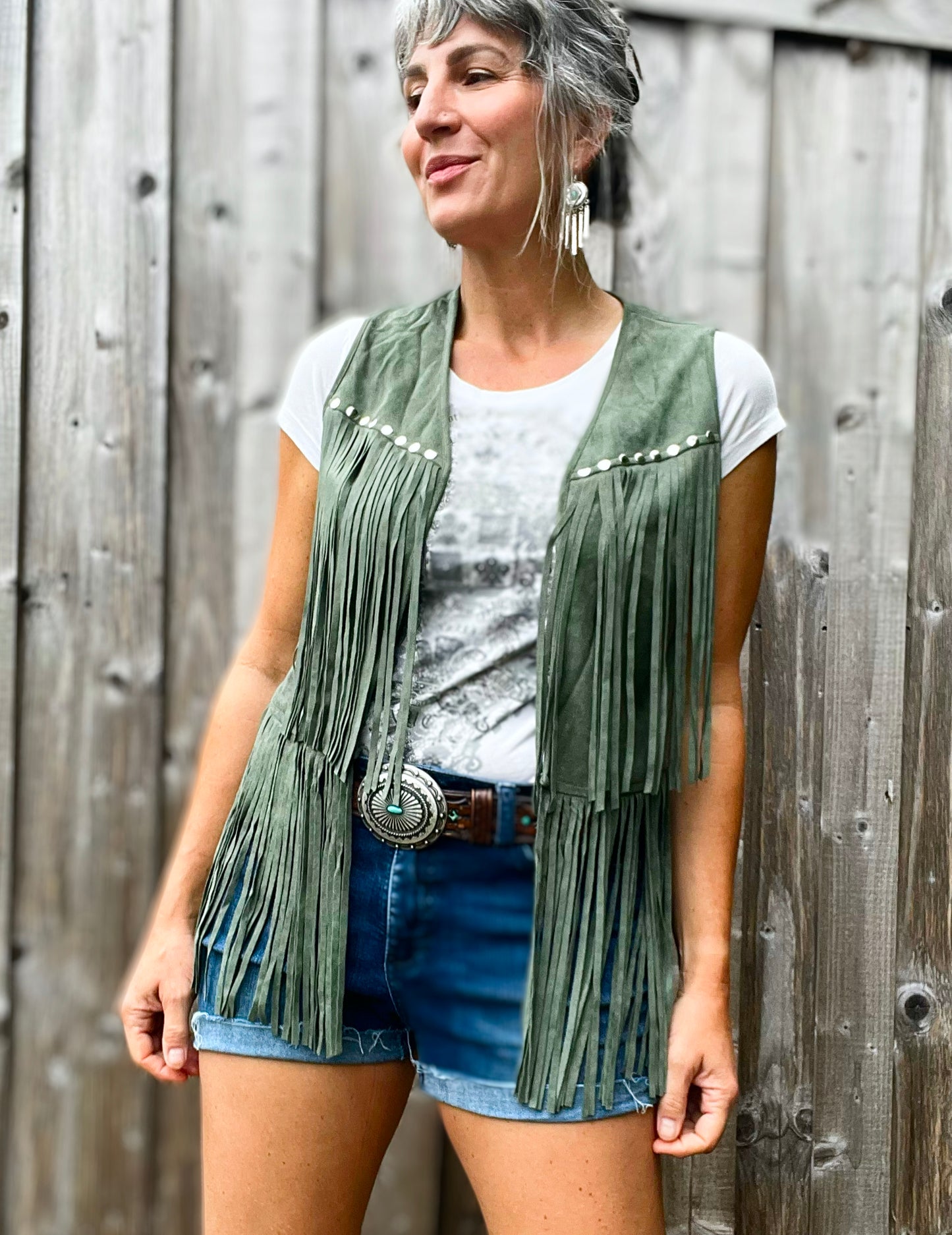 A woman wearing a green fringed Western style vest, standing against a wood outbuilding.