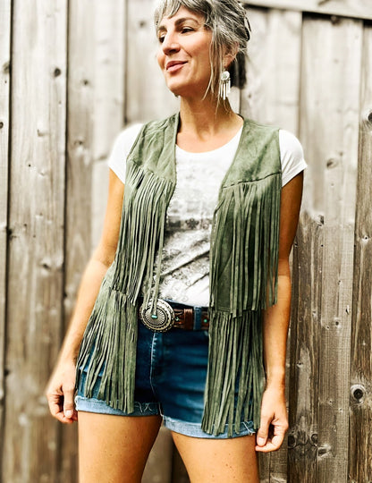 A woman wearing a moss green fringed vest with a Western style belt and cutoff shorts.