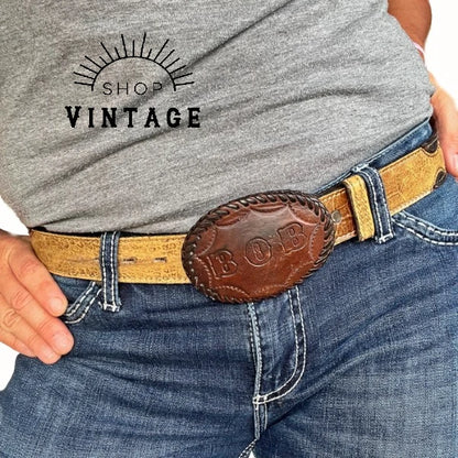 A close up of a person wearing a Western belt with a leather covered buckle that reads "Bob" in tooled leather lettering