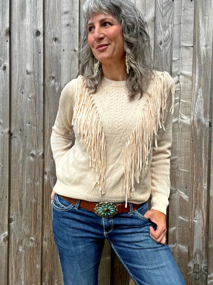 A woman standing against a wooden barn wall wearing a cream colored pullover Western fringe sweater.