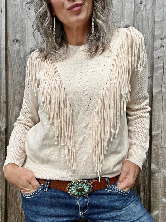 A woman standing next to a wooden barn wall wearing an off white fringed pullover sweater