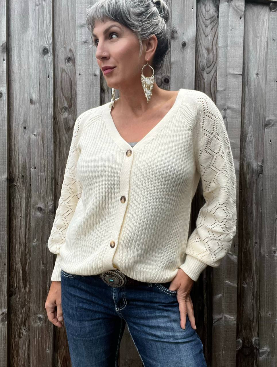 A woman standing against a barn wall wearing the Lauren button down cardigan in cream, paired with denim, beaded native earrings and a Western belt.