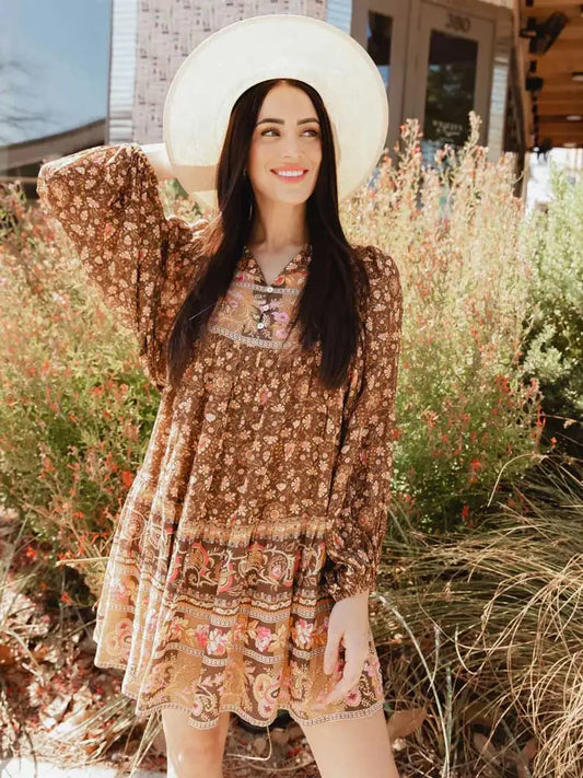 A woman standing against a desert backdrop wearing a brown boho inspired, loose fitting minidress with long sleeves and a white Western fedora.